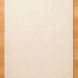May include: A rectangular, off-white rug with a textured, woven surface. The rug is placed on a light brown hardwood floor. The rug's design features a repeating pattern of small, raised dots, creating a tactile and visually interesting surface.