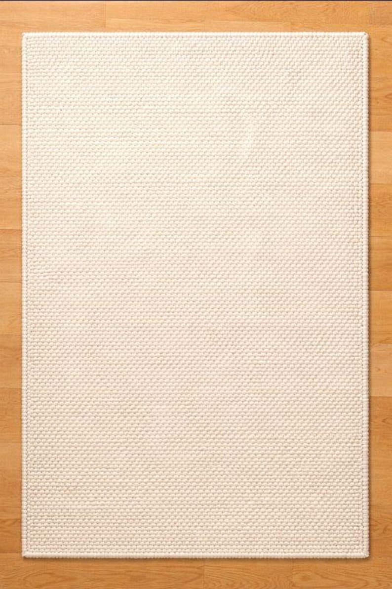 May include: A rectangular, off-white rug with a textured, woven surface. The rug is placed on a light brown hardwood floor. The rug's design features a repeating pattern of small, raised dots, creating a tactile and visually interesting surface.