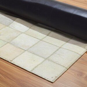 May include: A rolled-up area rug with a patchwork design. The rug features squares in shades of cream, beige, and light green, stitched together with visible seams. The backing is a dark, rolled material, possibly leather.