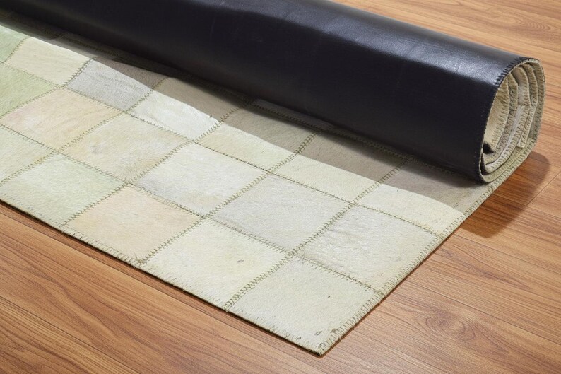 May include: A rolled-up area rug with a patchwork design. The rug features squares in shades of cream, beige, and light green, stitched together with visible seams. The backing is a dark, rolled material, possibly leather.