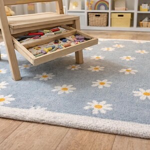 May include: A light blue rug featuring a daisy pattern, a wooden step stool with a drawer containing crayons, and a wooden toy. The rug has a white border and is on a wooden floor. A stuffed teddy bear is on the rug.