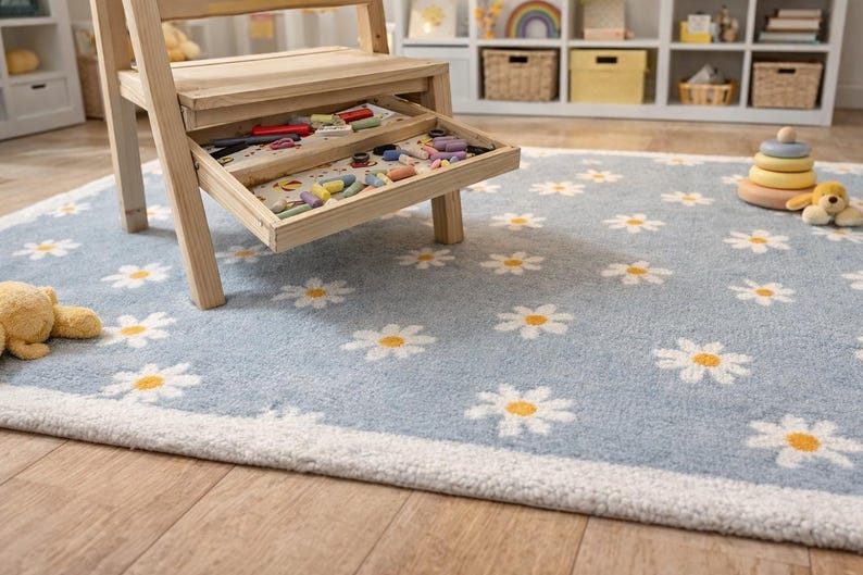 May include: A light blue rug featuring a daisy pattern, a wooden step stool with a drawer containing crayons, and a wooden toy. The rug has a white border and is on a wooden floor. A stuffed teddy bear is on the rug.