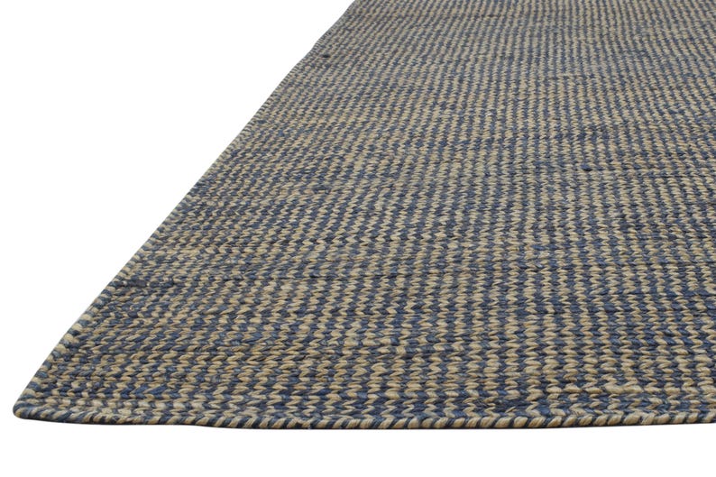 Puede incluir: Una alfombra rectangular con un patr&oacute;n tejido de rayas azules y beige alternas. La alfombra est&aacute; hecha de fibras naturales y tiene un aspecto texturizado. El borde de la alfombra es visible, mostrando un borde tejido apretado.