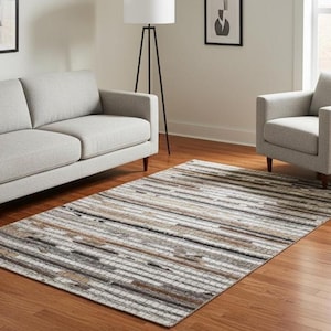 May include: Rectangular area rug with a striped pattern in shades of white, gray, and brown. The rug is placed on a hardwood floor in a living room setting, with a gray sofa and armchair.