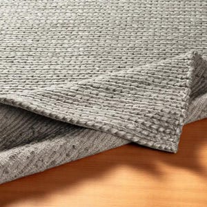 May include: A gray woven rug with a textured surface. The rug is folded over, revealing the underside. The rug's weave creates a pattern of raised, horizontal lines. The rug is made of a soft, durable material.