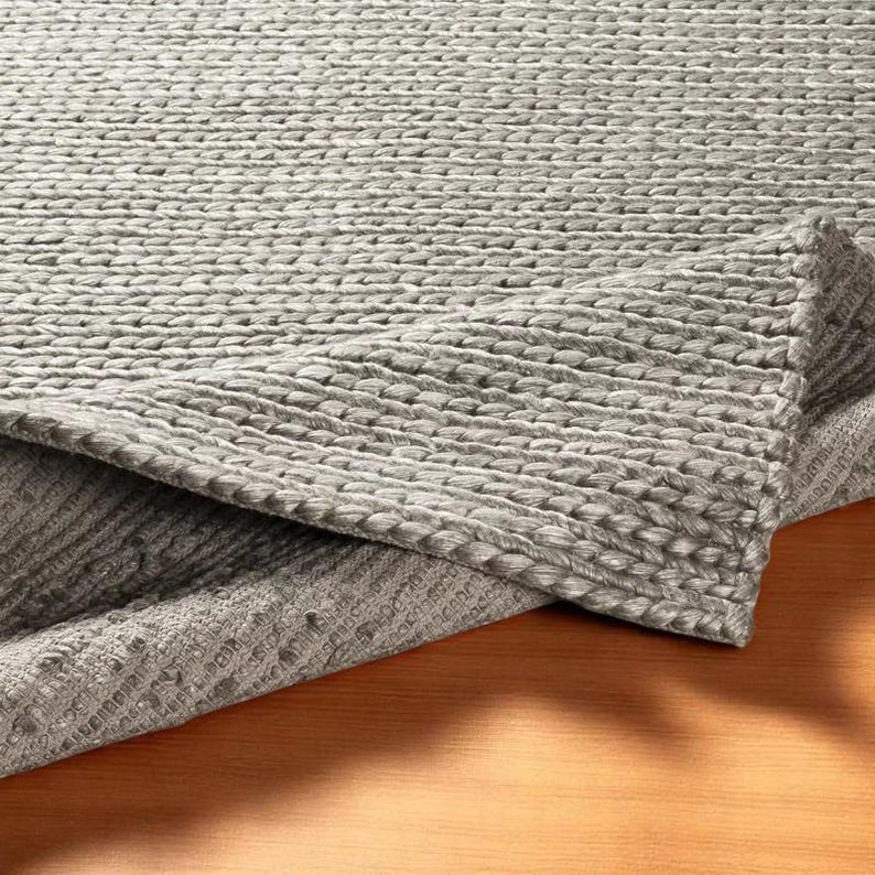 May include: A gray woven rug with a textured surface. The rug is folded over, revealing the underside. The rug's weave creates a pattern of raised, horizontal lines. The rug is made of a soft, durable material.