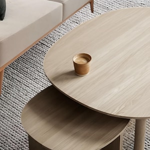 May include: A light wood oval coffee table set with a smaller table nested underneath. A small cup of coffee sits on the top table. The tables are in front of a beige sofa and a gray rug.