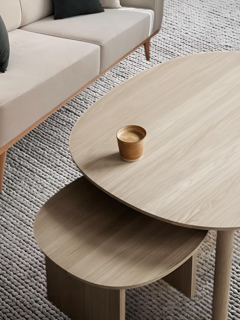 May include: A light wood oval coffee table set with a smaller table nested underneath. A small cup of coffee sits on the top table. The tables are in front of a beige sofa and a gray rug.