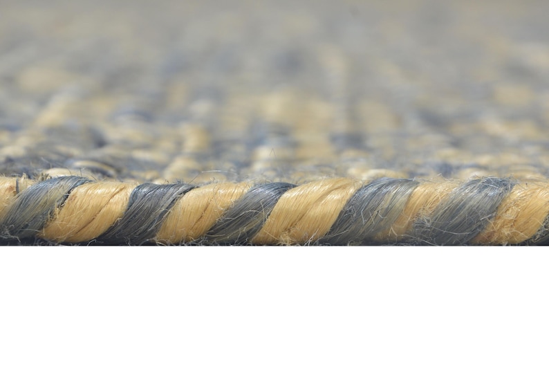 Puede incluir: Primer plano de una alfombra tejida con superficie texturizada. La alfombra presenta un patr&oacute;n de fibras azul-gris y beige alternadas, creando una apariencia trenzada. El borde de la alfombra es visible, mostrando el grosor y la construcci&oacute;n.