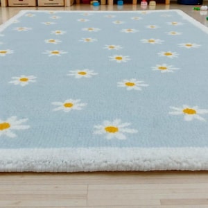 May include: A rectangular, pale blue rug featuring a white border and a pattern of white daisies with yellow centres. The rug is placed on a light-coloured wooden floor. The rug is made of a soft, plush material.