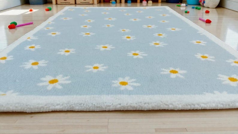 May include: A rectangular, pale blue rug featuring a white border and a pattern of white daisies with yellow centres. The rug is placed on a light-coloured wooden floor. The rug is made of a soft, plush material.