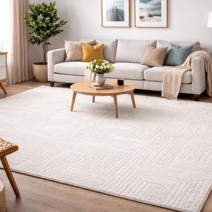 May include: A living room scene featuring a large, off-white geometric patterned rug. A light-colored sofa with decorative pillows sits against a wall with framed artwork. A round wooden coffee table and side table are in the foreground, with plants and lamps adding warmth.