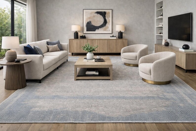 May include: A modern living room featuring a light-colored sofa with accent pillows, two swivel chairs, and a wooden coffee table. The room has a large area rug with a blue border and a neutral color palette. Two lamps sit on a wooden console.