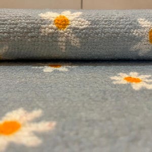 May include: A rolled-up, pale blue, plush fabric with a repeating pattern of white daisies with orange centres. The soft material is suitable for rugs, blankets, or other home decor items.