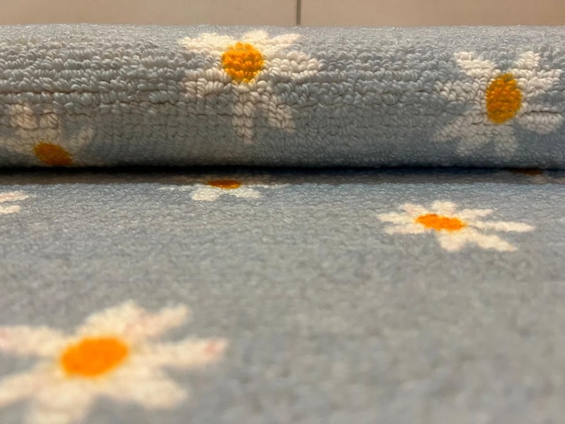 May include: A rolled-up, pale blue, plush fabric with a repeating pattern of white daisies with orange centres. The soft material is suitable for rugs, blankets, or other home decor items.