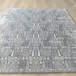 May include: A rectangular gray rug with a detailed floral pattern. The rug features a repeating design of stylized trees and leaves in shades of gray and white. The rug is placed on a light-colored wooden floor.