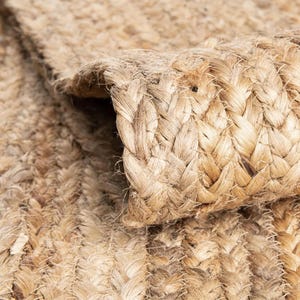 May include: Close-up of a natural fiber rug with a braided texture. The rug is a light tan color, with visible variations in tone. The edges of the rug are slightly curled, revealing the woven construction. The rug appears to be made of jute or a similar material.