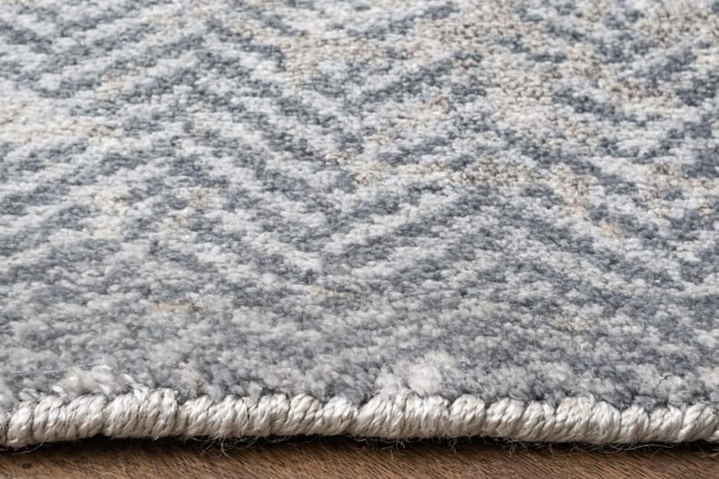 May include: Close-up of a textured area rug with a repeating chevron pattern. The rug features a blend of gray, off-white, and beige tones, creating a neutral color palette. The edges are finished with a light-colored border.