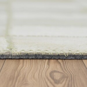 May include: Close-up of a light-colored rug with a stitched border. The rug has a grid pattern and a soft, textured appearance. The bottom edge of the rug is visible, showing the stitching and the backing material. The rug is on a wooden surface.