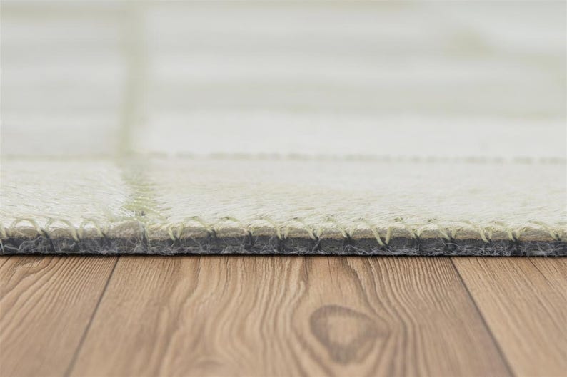 May include: Close-up of a light-colored rug with a stitched border. The rug has a grid pattern and a soft, textured appearance. The bottom edge of the rug is visible, showing the stitching and the backing material. The rug is on a wooden surface.