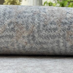 May include: Rolled-up area rug with a textured surface. The rug features a pattern of gray, blue, and beige hues. The fibers create a soft, plush appearance, suitable for home decor.