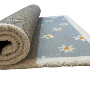 May include: Rolled-up rug with a light blue field featuring white daisies with yellow centres. The rug has a white border and a beige backing. The rug is partially rolled, revealing the underside and the daisy pattern.