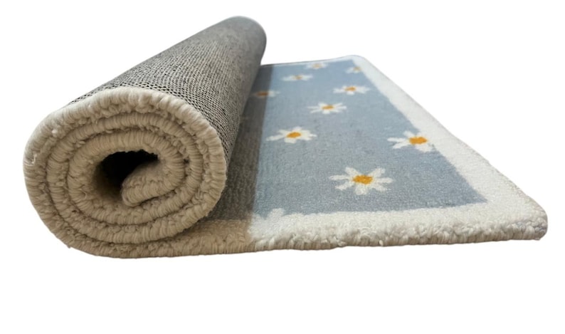 May include: Rolled-up rug with a light blue field featuring white daisies with yellow centres. The rug has a white border and a beige backing. The rug is partially rolled, revealing the underside and the daisy pattern.