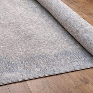 May include: A rolled-up, rectangular area rug with a faded, distressed appearance. The rug features a light gray and blue color palette with a subtle geometric border design. The rug is partially unrolled on a wooden floor.