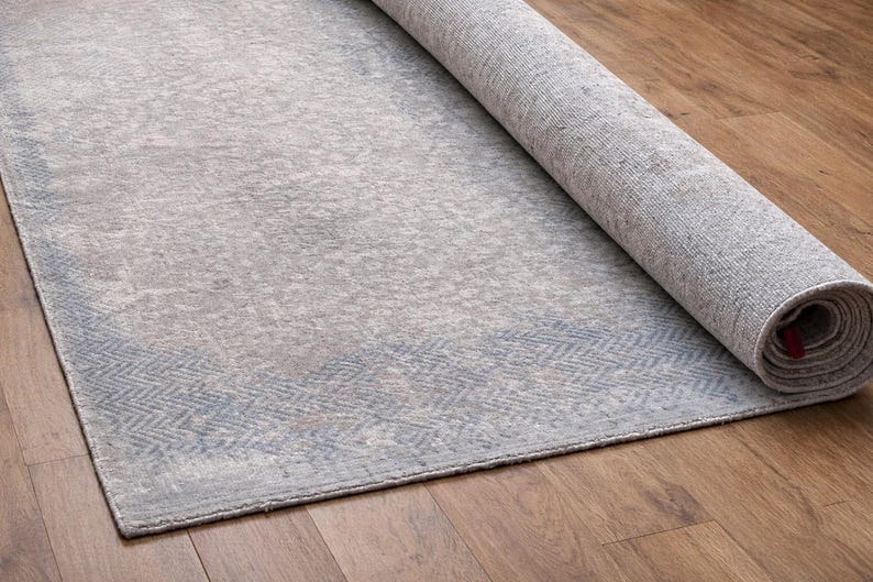 May include: A rolled-up, rectangular area rug with a faded, distressed appearance. The rug features a light gray and blue color palette with a subtle geometric border design. The rug is partially unrolled on a wooden floor.