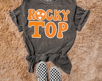 Puede incluir: Camiseta gris oscuro con las palabras "ROCKY TOP" en letras de bloque naranja con un contorno blanco. La palabra "ROCKY" tiene tres estrellas blancas. La camiseta se combina con zapatos sin cordones a cuadros en blanco y negro.