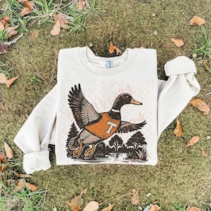 May include: Cream-colored sweatshirt featuring a graphic of a duck wearing an orange vest with a white "T" on it. The duck is flying over a body of water with trees in the background. The sweatshirt has long sleeves.