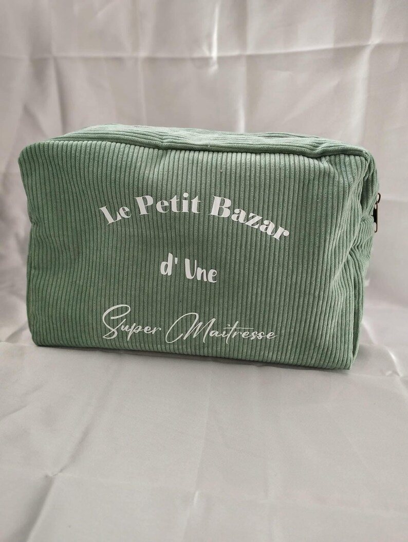 Pu&ograve; includere: Un beauty case rettangolare in velluto a coste verde salvia. La borsa presenta il testo "Le Petit Bazar d'Une Super Ma&icirc;tresse" in scrittura bianca. La borsa ha una chiusura con cerniera e una texture a coste.