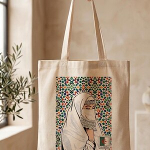 May include: A beige canvas tote bag featuring a graphic print of a person wearing a white head covering and face veil. The background of the print displays a colorful geometric mosaic pattern, and a small stamp-like icon reads ALGERIA.