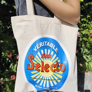 May include: A beige canvas tote bag with cream-colored handles. The bag features a large, circular graphic with the words "Veritable Soda Selecto" in red and white against a blue background with yellow rays. The text "Boisson rafraîchissante sans alcool" is also visible.