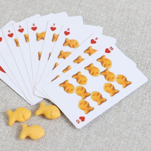 May include: A deck of playing cards with heart suits and goldfish cracker designs. The cards are fanned out, with some goldfish crackers scattered around them. The cards show the numbers 4 through 10.