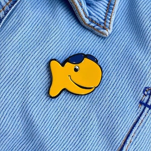 Könnte beinhalten: Ein gelber Emaille-Pin in Form eines lächelnden Fisches mit blauer Sonnenbrille. Der Pin hat eine schwarze Umrandung und ist an einer hellblauen Jeansjacke befestigt.
