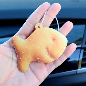 May include: A yellow fish-shaped car air freshener hanging from a string. The air freshener is made to look like a cracker.
