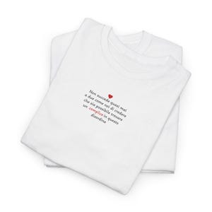 May include: Two white t-shirts are stacked, with the top one featuring text in Italian and a small red heart. The text reads, "Non succede quasi mai a due come noi di credere che sia possibile trovare un complice in questo disordine."