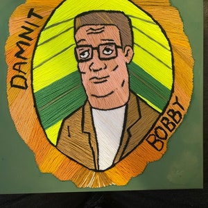May include: A green background with a yellow, green, and orange oval frame. Inside the frame is a portrait of a man with brown hair, glasses, and a brown jacket. The text "DAMNIT BOBBY" is written around the frame.
