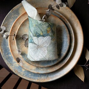 Pode incluir: Uma mesa rústica com pratos em camadas em tons de azul e bege. Um porta-guardanapos floral com um desenho de flor branca está sobre os pratos. Ramos de eucalipto e uma vela em um suporte de cobre completam o conjunto.