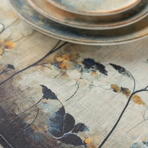 May include: A beige linen table runner with a floral design in shades of blue, brown, and yellow. The runner features a subtle textured pattern and is accented with a set of stacked ceramic plates.