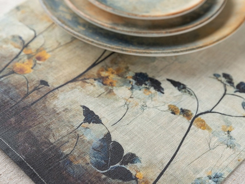 May include: A beige linen table runner with a floral design in shades of blue, brown, and yellow. The runner features a subtle textured pattern and is accented with a set of stacked ceramic plates.