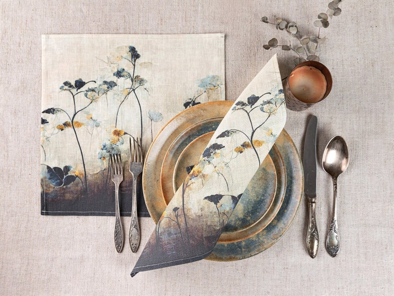 May include: A set of two linen placemats and napkins with a floral design in shades of blue, yellow, and brown. The placemats and napkins are folded and arranged on a beige linen tablecloth with silver cutlery and a ceramic plate.