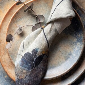 May include: A set of three ceramic plates with a blue and brown glaze. A white linen napkin with a floral pattern is folded and placed on top of the plates.