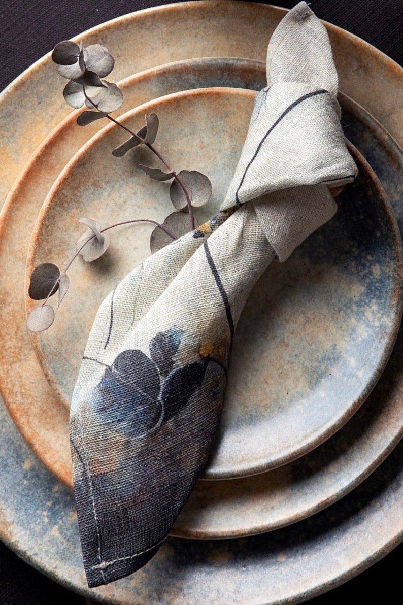 May include: A set of three ceramic plates with a blue and brown glaze. A white linen napkin with a floral pattern is folded and placed on top of the plates.