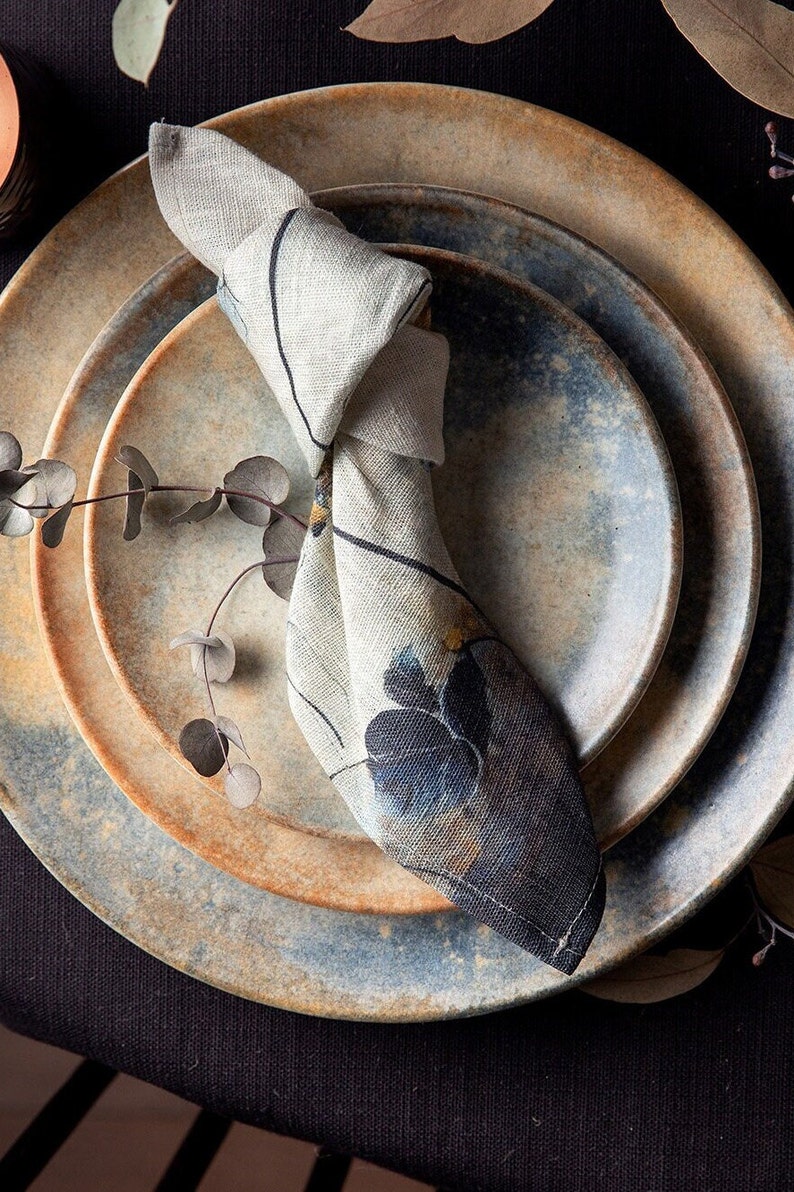 May include: A set of three ceramic plates with a blue and brown speckled glaze. A patterned linen napkin is placed on top of the plates.
