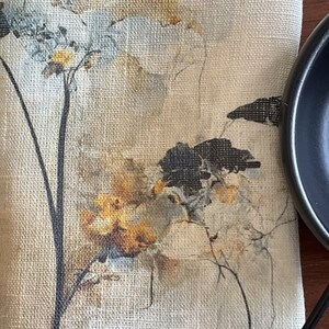 May include: A beige linen fabric with a floral design printed on it. The design features black silhouettes of birds and brown and white watercolor flowers.