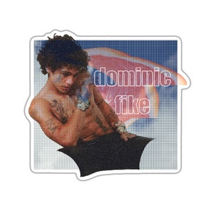 Puede incluir: Una pegatina con una foto de un hombre sin camisa con cabello oscuro y tatuajes. El hombre est&aacute; sentado con los brazos cruzados y mirando hacia abajo. El fondo es un patr&oacute;n de cuadr&iacute;cula azul y blanco con un gr&aacute;fico de fruta roja y naranja. El texto "dominic fike" est&aacute; escrito en letras blancas delineadas en el gr&aacute;fico de la fruta.