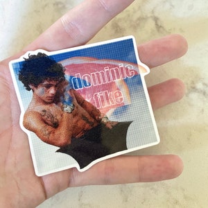 Puede incluir: Una pegatina con una foto de Dominic Fike y el texto "dominic fike" en letras blancas sobre un fondo azul y rosa.