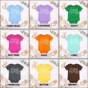 Puede incluir: Nueve bodies de beb&eacute; en varios colores: azul claro, lavanda, verde kelly, rosa intenso, dorado, chill, carb&oacute;n, amarillo mantequilla y marr&oacute;n. Cada body tiene el texto "PAPER HANGER DESIGNS".